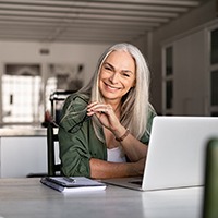 An older woman seated behind her computer and smiling after receiving her customized dental implants